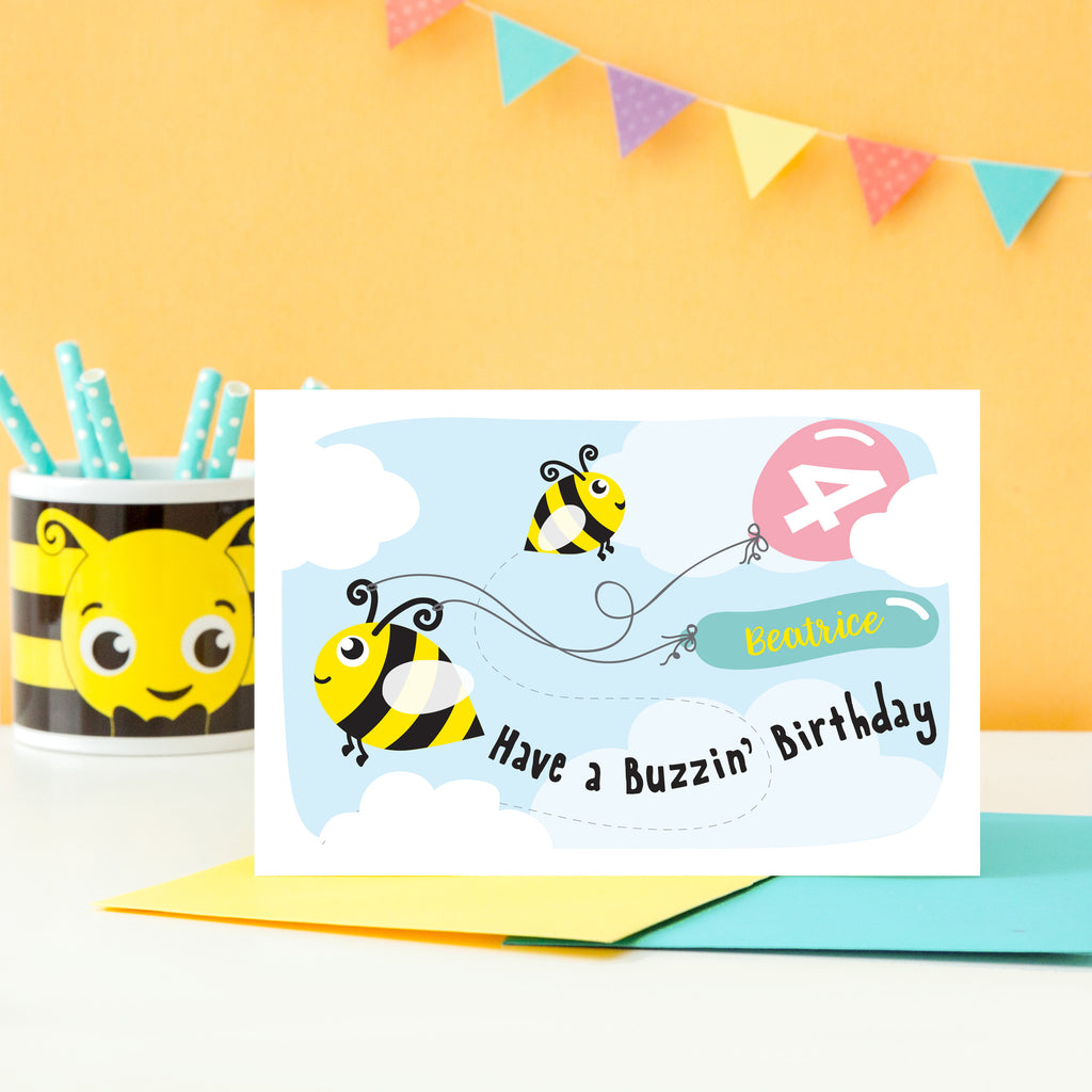 Cute personalised bee birthday card shows two bees in the sky with clouds pulling balloons with any name and age added. Shown here with Pink balloons.