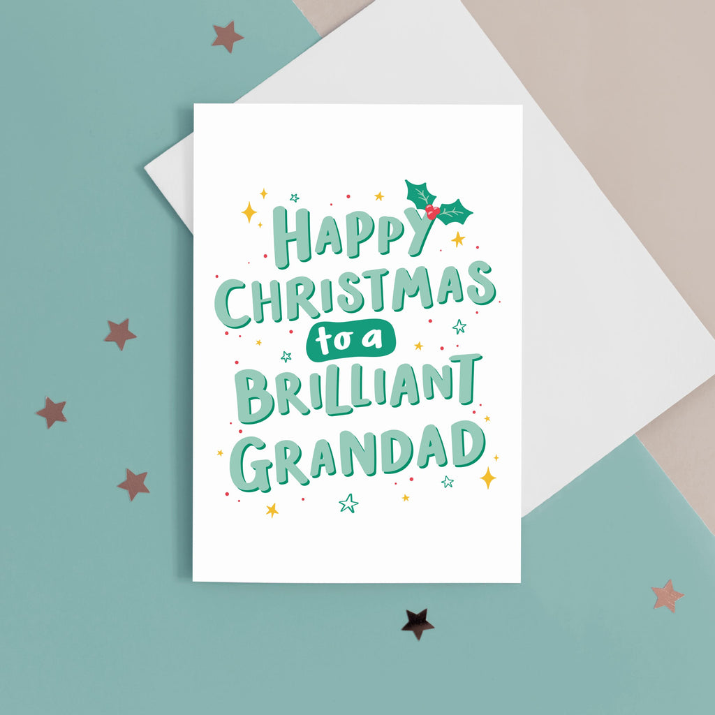 A hand lettered typographic card which reads 'Happy Christmas to a brilliant Grandad'. The card features mint green lettering with a dark green drop shadow, a sprig of holly and a collection of star flourishes around the letterring. The card can be personalised with Grandad's special name.