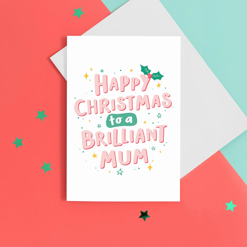 A hand lettered typographic card which reads 'Happy Christmas to a brilliant Mum'. The card features pink letterring with a red drop shadow, a sprig of holly and a collection of star flourishes around the letterring. The card can be personalised with Mum or Mummy.
