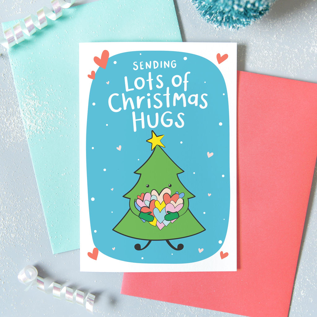 Sending lots of Christmas hugs. A cute colourful Christmas card with a little Christmas tree hugging a bunch of colourful love hearts. Against a blue background with snow and hearts with a yellow star above the tree.