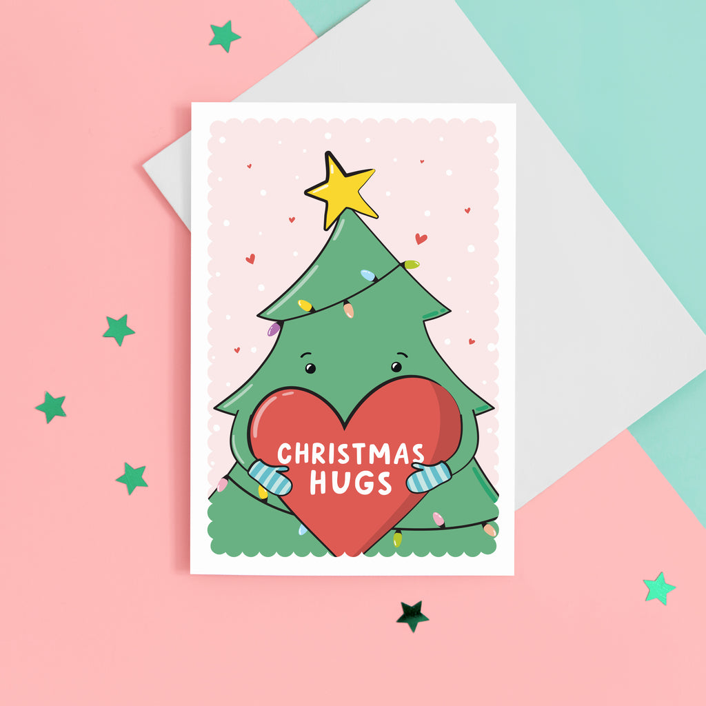 A cute Christmas card featuring a cartoon Christmas tree hugging a large love heart. The love heart reads, "Christmas hugs". A yellow star is on the top of the tree set against a pink background with love hearts.