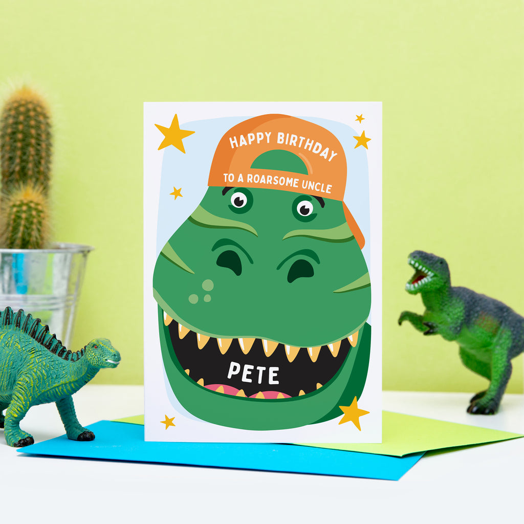 A bold and fun birthday card featuring the face of a dinosaur with a big grin, wearing a bright orange baseball hat. The card reads Happy Birthday to a roarsome Uncle with space to personalise with a name.