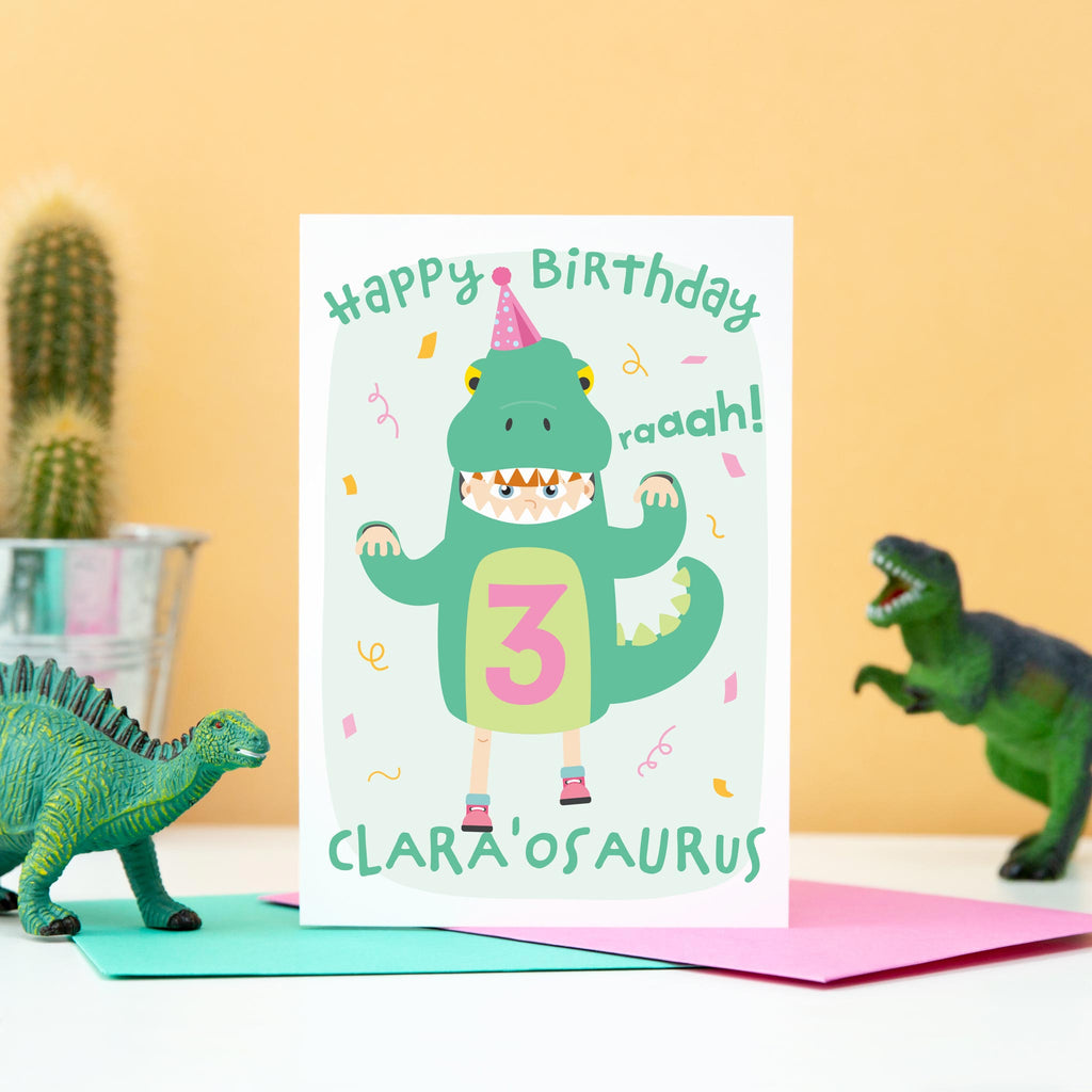 Birthday card with cute girl dressed in a dinosaur costume. Personalised for any name and age, as well age eye colour, skin colour and hair colour. Shown age three.