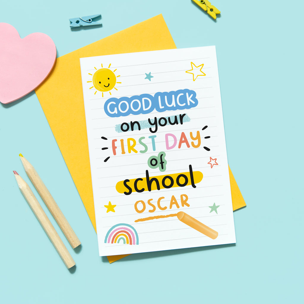 A bright and colourful back to school card which reads, 'good luck on your first day of school' with the name of the recipient underneath. The card features hand drawn text with small graphics of a sunshine, rainbow and stars.