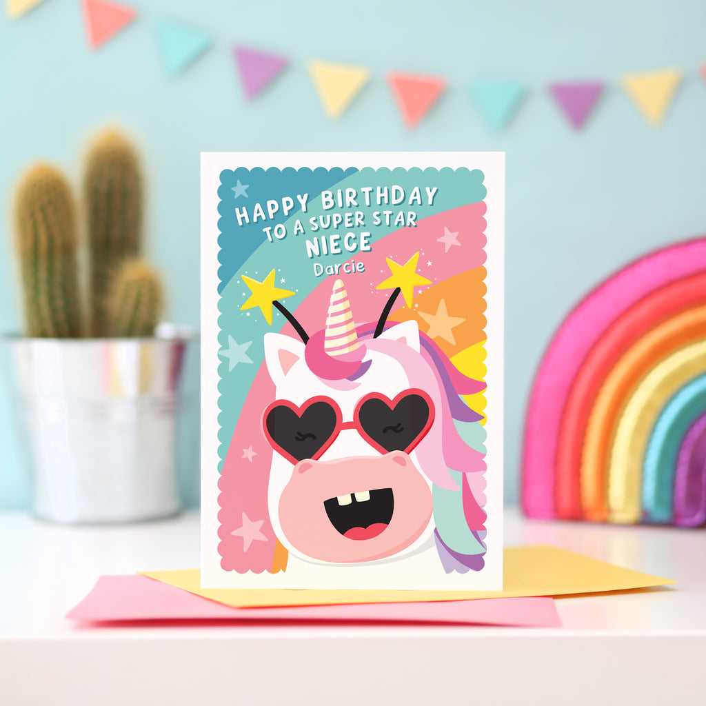 A fun and colourful birthday card featuring a happy unicorn face, wearing heart glasses and a star headband. The card reads ‘Happy Birthday to a super star Niece’ but can be personalised for any recipient and has space to add the recipients name.