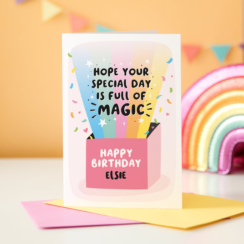 A colourful birthday card featuring a bright rainbow and confetti exploding out of a gift box. The rainbow features text in front saying 'hope your special day is full of magic' and the box says 'Happy Birthday' with a personalised name underneath.