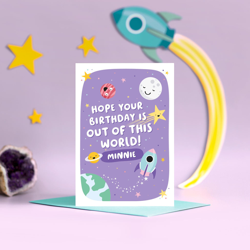 A cute and colourful card with a space theme, featuring happy planets, stars and a rocket with a purple background. The wording on the card says hope your birthday is out of this world with a space for personalising with a name.