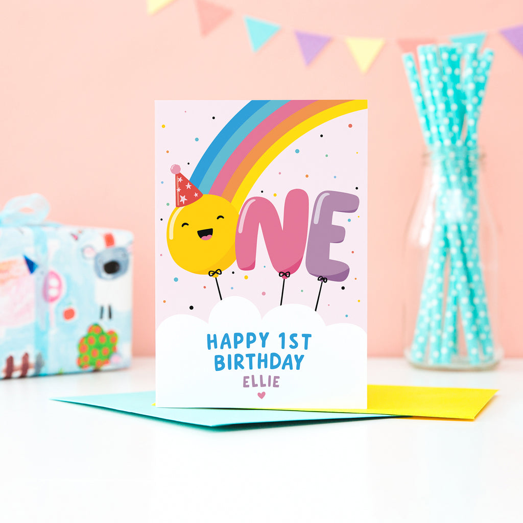 A bold and colourful card featuring a rainbow and the word 'One' designed as balloons. The 'O' balloon has a happy face and wears a party hat. The card has a white cloud at the bottom with text which reads, ' Happy 1st Birthday’, with space to personalise with a name.