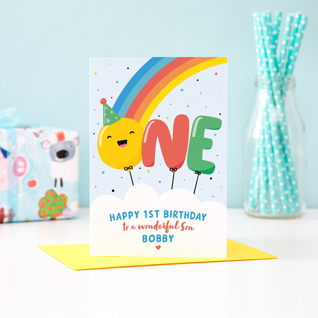 A bold and colourful card featuring a rainbow and the word 'One' designed as balloons. The 'O' balloon has a happy face and wears a party hat. The card has a white cloud at the bottom with text which reads, ' Happy 1st Birthday to a wonderful Son', with space to personalise with a name.