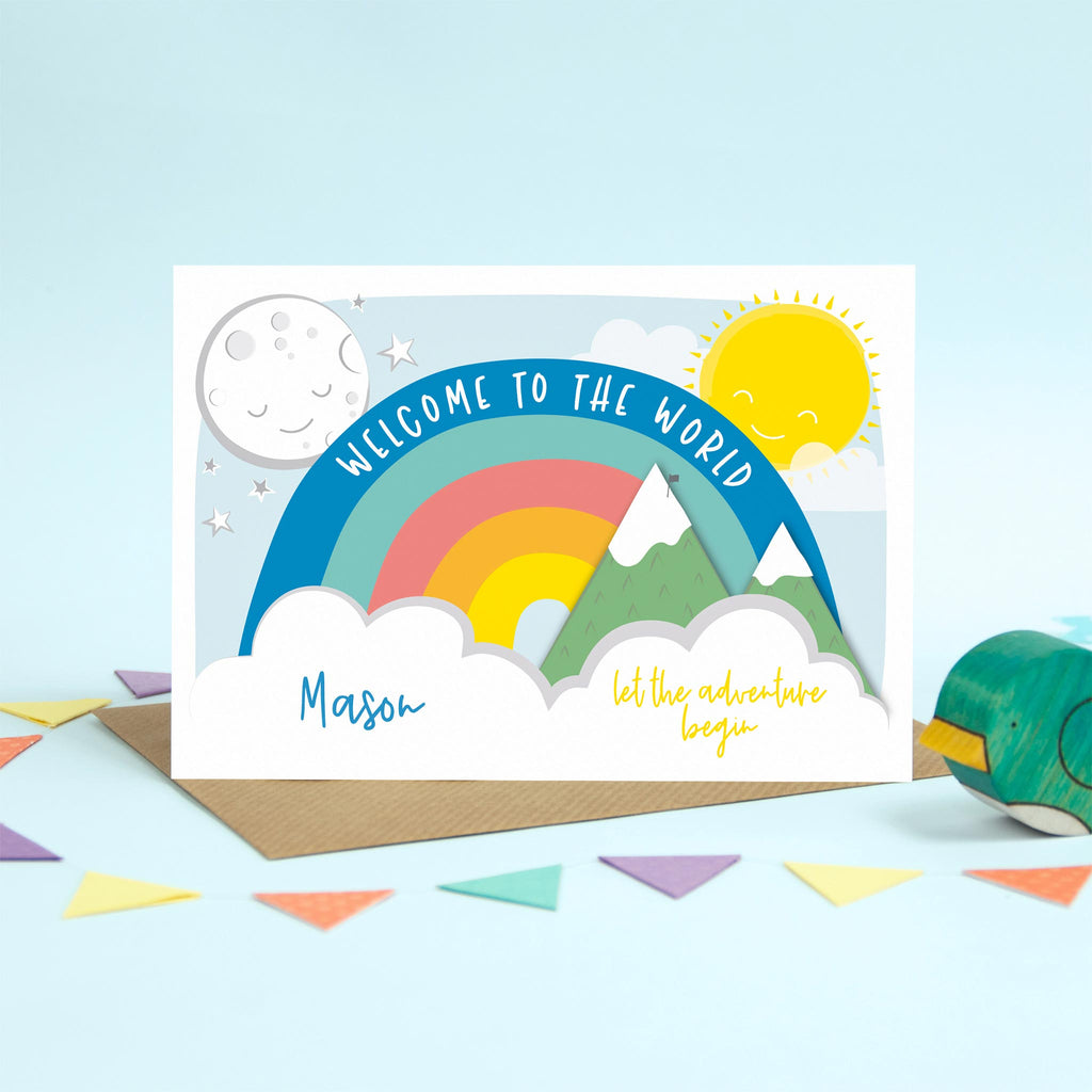 Blue writing on this colourful personalised new baby card with rainbow, sun, moon, mountains and clouds.