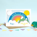 Load image into Gallery viewer, Blue writing on this colourful personalised new baby card with rainbow, sun, moon, mountains and clouds.
