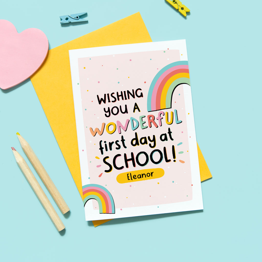 A colourful back to school card featuring rainbows on a light pink background with small colourful dots. The card reads, 'wishing you a wonderful first day at school', with a space underneath to personalise with the recipients name.