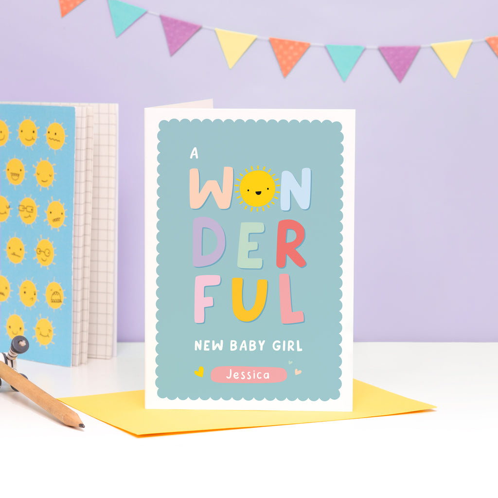 A colourful card featuring the words 'a wonderful new baby girl’ with space to personalise with a name. The card has a turquoise background and the word 'wonderful' has bright colours with a smiley sunshine face for the letter 'o'.