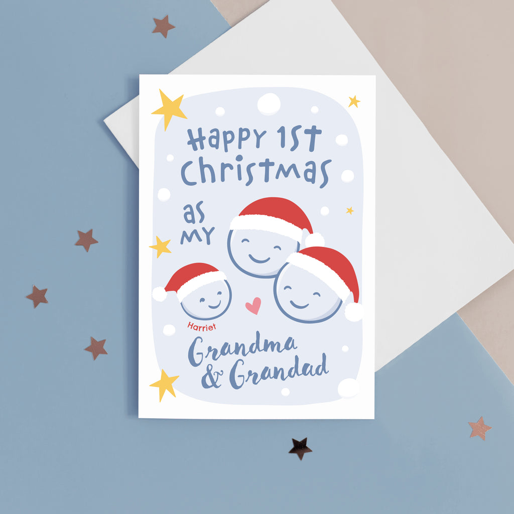 A card with a light blue background featuring stars and snow, with the words Happy 1st Christmas as my Grandma & Grandad. The card also features smiley faces for baby and Grandparents, wearing santa hats. The card can be personalised with the baby's name.