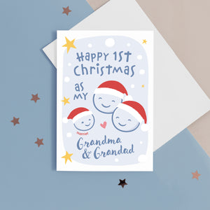 A card with a light blue background featuring stars and snow, with the words Happy 1st Christmas as my Grandma & Grandad. The card also features smiley faces for baby and Grandparents, wearing santa hats. The card can be personalised with the baby's name.