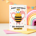 Load image into Gallery viewer, Happy birthday to a bee-rilliant Grandma. A cute and punny card featuring a happy bee wearing a party hat to represent Grandma, with a pink heart in the background and a sprinkle of confetti.
