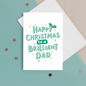 A hand lettered typographic card which reads 'Happy Christmas to a brilliant Dad'. The card features mint green lettering with a dark green drop shadow, a sprig of holly and a collection of star flourishes around the letterring. The card can be personalised with Dad or Daddy.