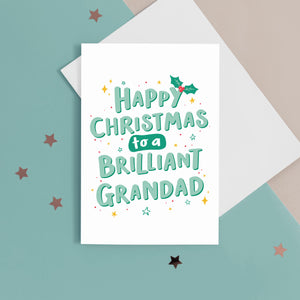 A hand lettered typographic card which reads 'Happy Christmas to a brilliant Grandad'. The card features mint green lettering with a dark green drop shadow, a sprig of holly and a collection of star flourishes around the letterring. The card can be personalised with Grandad's special name.