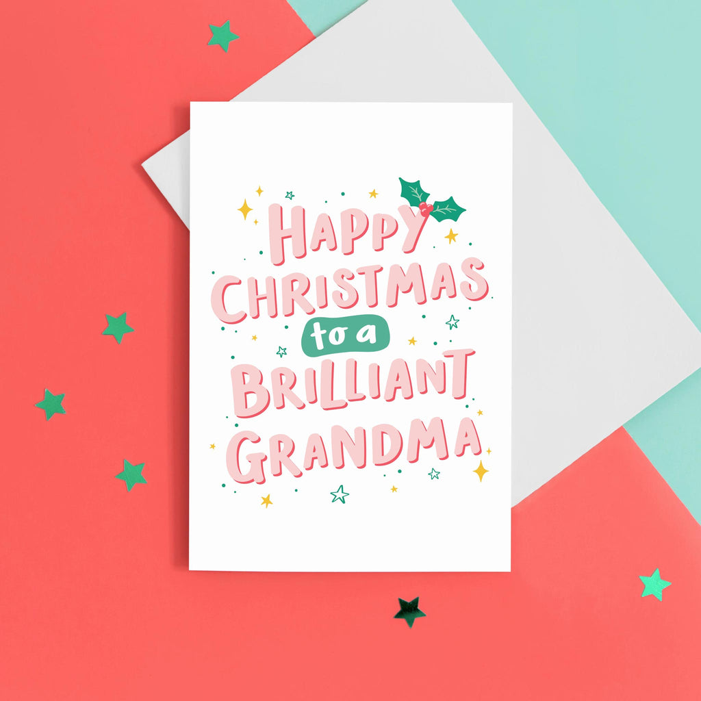 A hand lettered typographic card which reads 'Happy Christmas to a brilliant Grandma'. The card features pink letterring with a red drop shadow, a sprig of holly and a collection of star flourishes around the letterring.