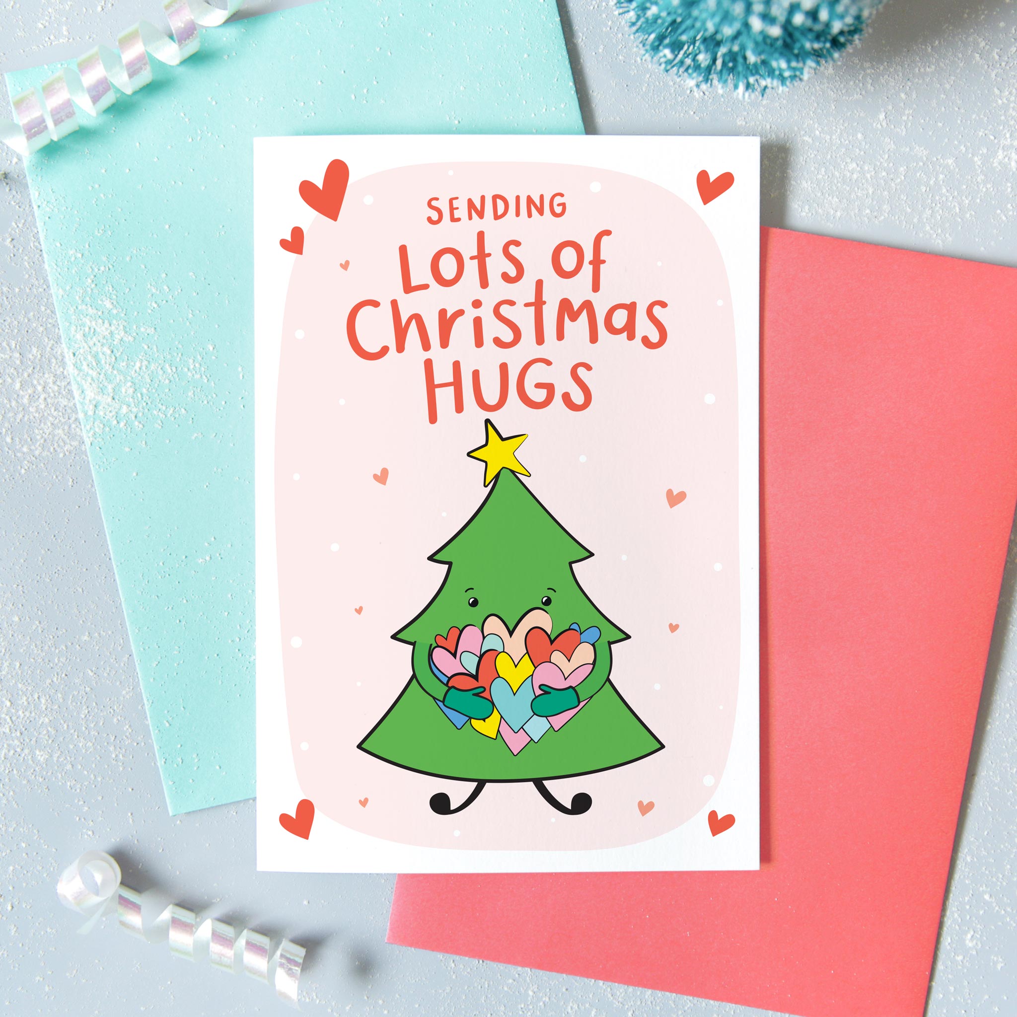 Sending lots of Christmas hugs. A cute colourful Christmas card with a little Christmas tree hugging a bunch of colourful love hearts. Against a pink background with snow and hearts with a yellow star above the tree.