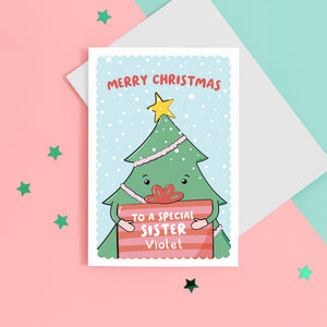 A cute Christmas card with a blue background, featuring a Christmas tree holding a present with a sprinkling of snow. The card reads Merry Christmas to a special Sister with space to personalise with a name.