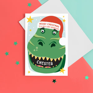 A bold and fun Christmas card featuring the face of a dinosaur with a big grin, wearing a red santas hat. The card reads Merry Christmas to a brilliant Grandson with space to personalise with your Grandson's name.