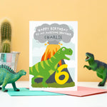 Load image into Gallery viewer, Dinosaur happy birthday card featuring t-rex and an erupting volcano. A personalised message and name can be added to the smoke above the volcano. shown age 6 to brother
