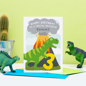 Dinosaur happy birthday card featuring t-rex and an erupting volcano. A personalised message and name can be added to the smoke above the volcano. shown age 3 to grandson