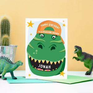 A bold and fun birthday card featuring the face of a dinosaur with a big grin, wearing a bright orange baseball hat. The card reads Happy Birthday to a roarsome Nephew.