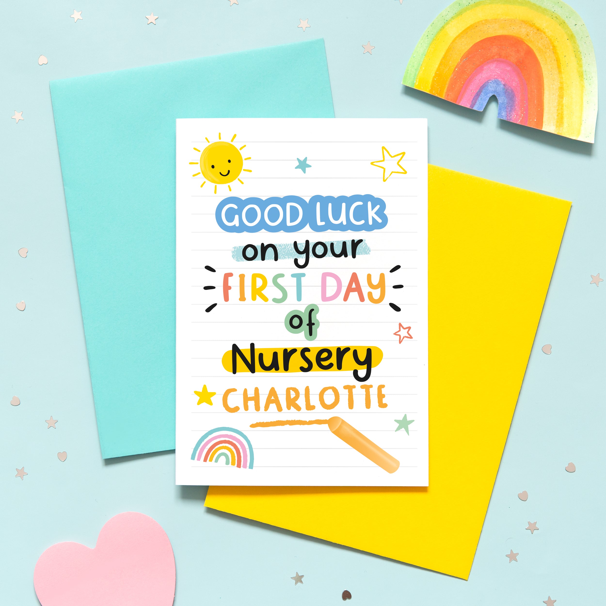 A bright and colourful back to school card which reads, 'good luck on your first day of nursery' with the name of the recipient underneath. The card features hand drawn text with small graphics of a sunshine, rainbow and stars.