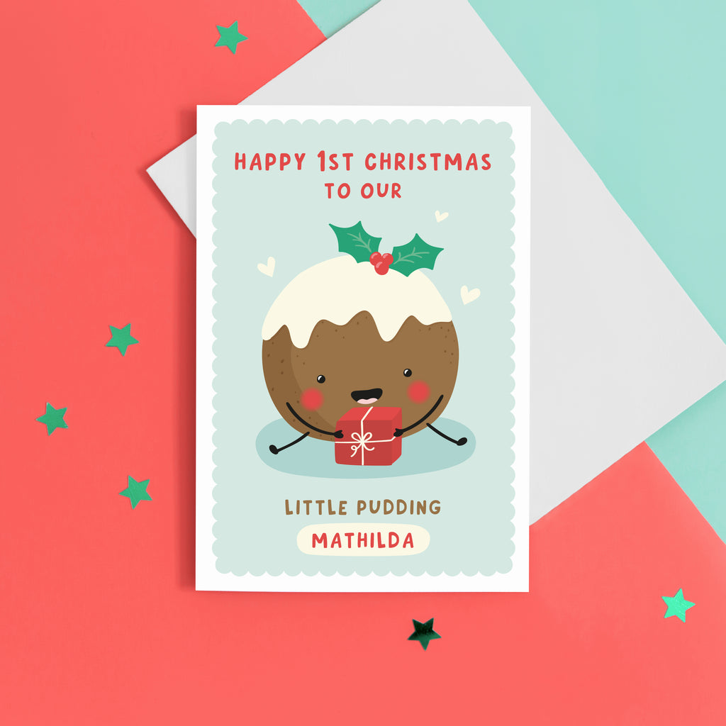 A super cute card featuring a happy little Christmas pudding illustration, holding a present. The card reads 'Happy 1st Christmas to our little pudding', with space to personalise with the baby's name.