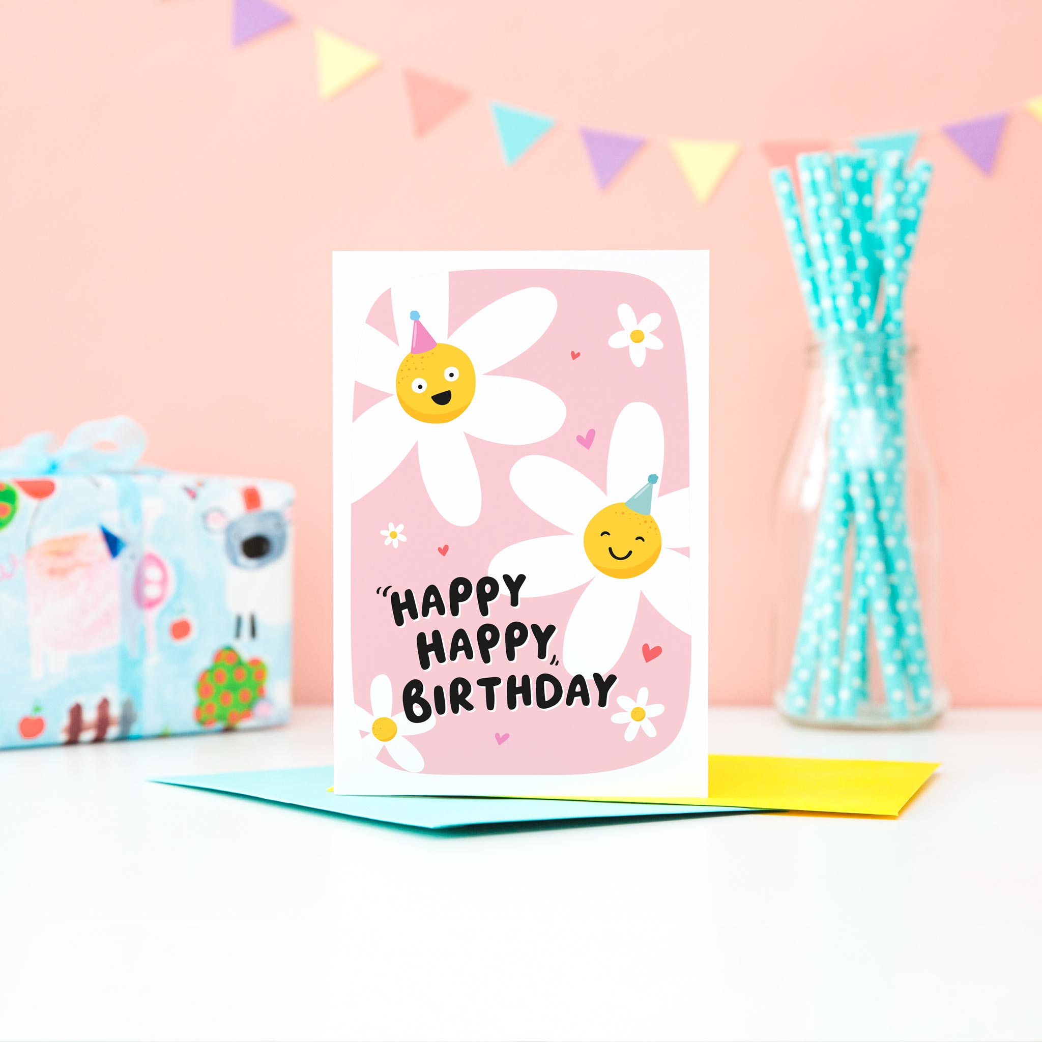 A cute card with a pink background featuring two happy daisies wearing party hats, a few hearts and the words happy happy birthday.