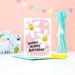 Load image into Gallery viewer, A cute card with a pink background featuring two happy daisies wearing party hats, a few hearts and the words happy happy birthday.
