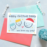 Load image into Gallery viewer, Happy Christmas Daddy, we love you lots. Cute Christmas card featuring 3 faces representing Daddy and 2 children. The smaller faces wear Christmas party hats and the larger face a santa hat. The card is personalised with the children&#39;s names and has a soft blue background.
