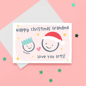 Happy Christmas Grandma, love you lots. Cute Christmas card featuring 2 faces representing Grandmother and Grandchild. The smaller face wears a Christmas party hat and a larger face a santa hat. The card is personalised with the child's name and has a soft pink background.