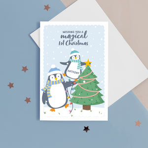 A cute Christmas card with a light blue background, featuring a baby penguin balancing on a big penguin whilst putting a star on the top of a Christmas tree. The card reads ‘Wishing you a magical 1st Christmas’. The card has space to place the baby’s name on the baby penguins belly.