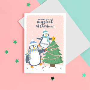 A cute Christmas card with a light peach background, featuring a baby penguin balancing on a big penguin whilst putting a star on the top of a Christmas tree. The card reads ‘Wishing you a magical 1st Christmas’. The card has space to place the baby’s name on the baby penguins belly.