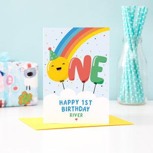 A bold and colourful card featuring a rainbow and the word 'One' designed as balloons. The 'O' balloon has a happy face and wears a party hat. The card has a white cloud at the bottom with text which reads, ' Happy 1st Birthday’, with space to personalise with a name.