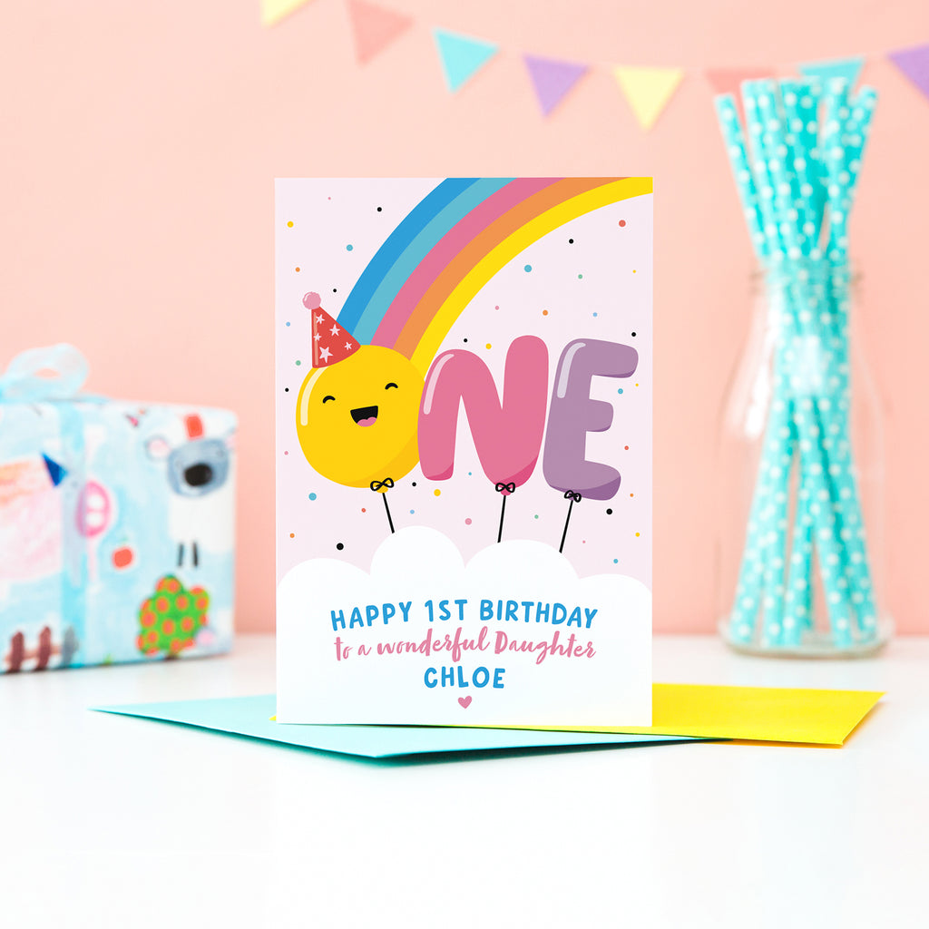 A bold and colourful card featuring a rainbow and the word 'One' designed as balloons. The 'O' balloon has a happy face and wears a party hat. The card has a white cloud at the bottom with text which reads, ' Happy 1st Birthday to a wonderful Daughter', with space to personalise with a name.