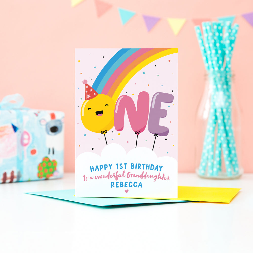 A bold and colourful card featuring a rainbow and the word 'One' designed as balloons. The 'O' balloon has a happy face and wears a party hat. The card has a white cloud at the bottom with text which reads, ' Happy 1st Birthday to a wonderful Granddaughter', with space to personalise with a name.