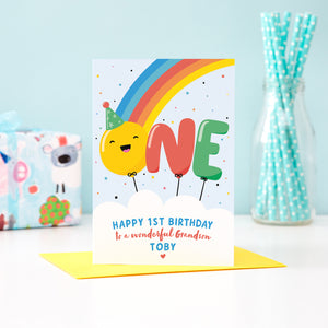 A bold and colourful card featuring a rainbow and the word 'One' designed as balloons. The 'O' balloon has a happy face and wears a party hat. The card has a white cloud at the bottom with text which reads, ' Happy 1st Birthday to a wonderful Grandson', with space to personalise with a name.