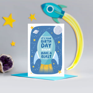 A bright and colourful birthday card featuring a rocket and a space scene with the  words, 'It's your birthday have a blast'. There is an option to personalise this card with a name.