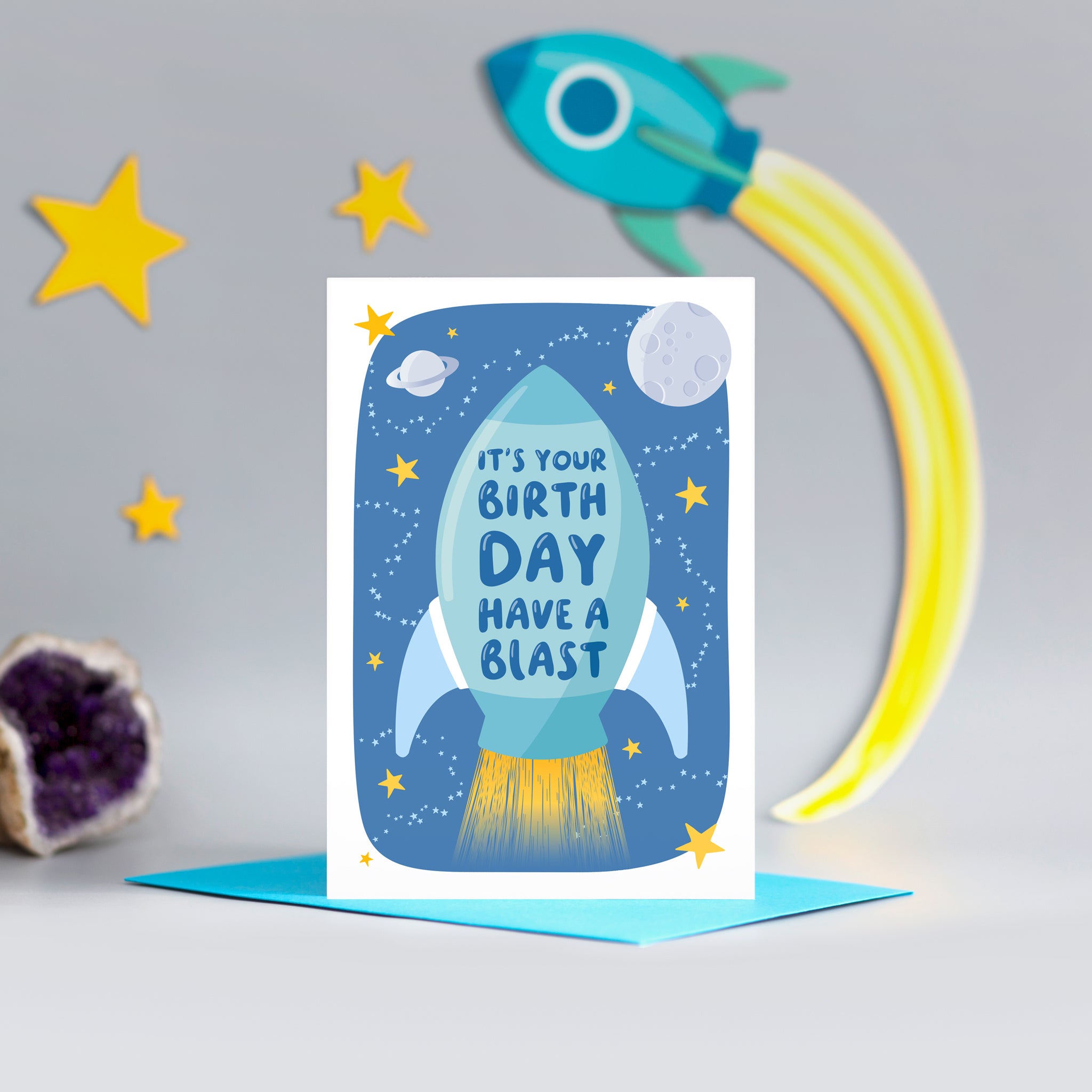 A bright and colourful birthday card featuring a rocket and a space scene with the words, 'It's your birthday have a blast'.