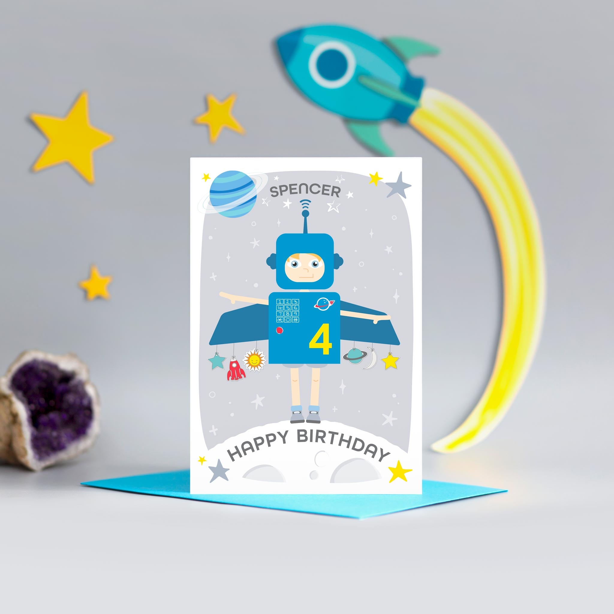 Space boy happy birthday card. This fun and colourful card shows a little boy dressed up as a blue space rocket with arms out. The card also features a moon, planets and stars. The card is personalised with any name and age.