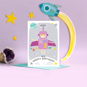 Space girl happy birthday card. This fun and colourful card shows a little girl dressed up as a purple space rocket with arms out. The card also features a moon, planets and stars. The card is personalised with any name and age.