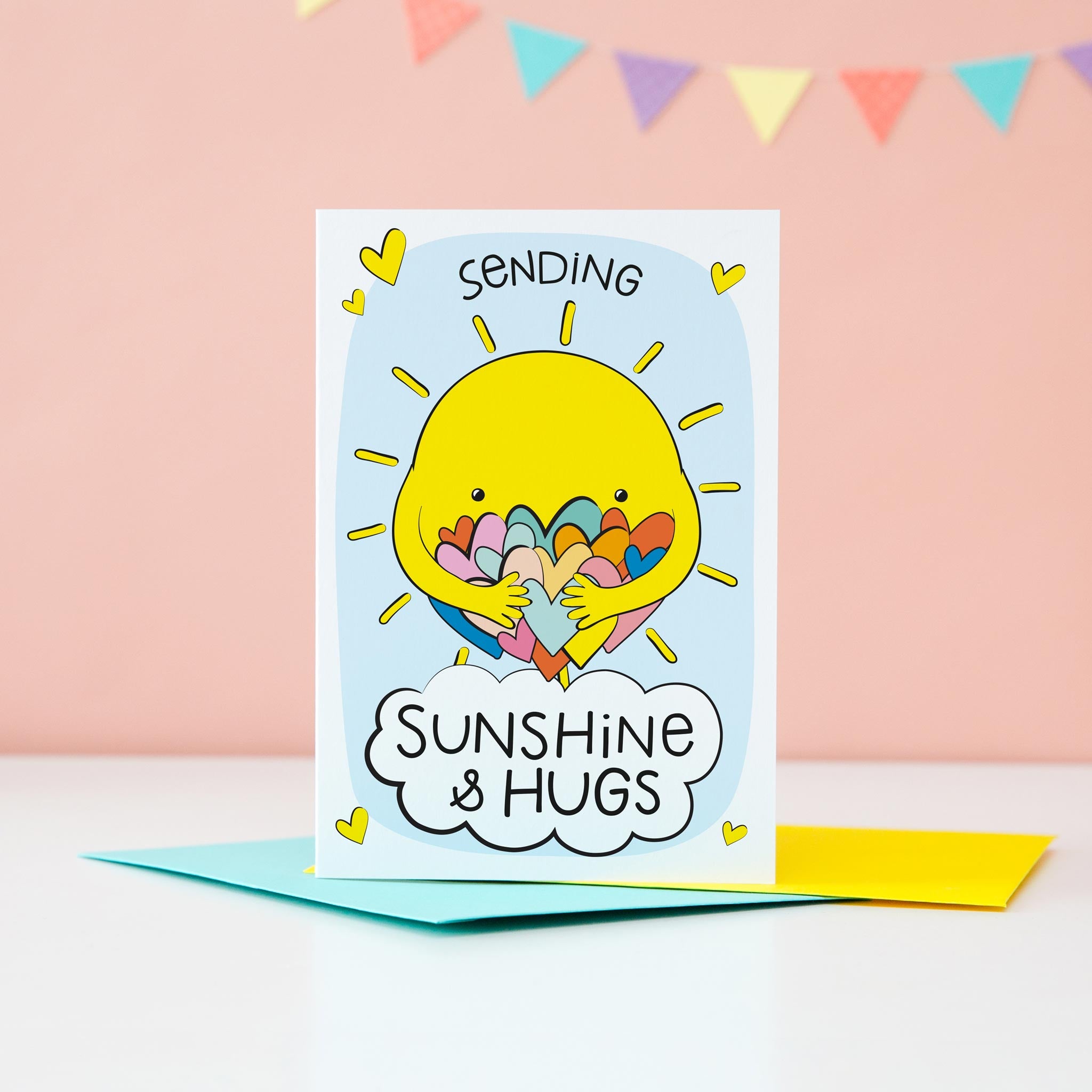 A bright and colourful cute card which features a shining sun hugging a large number of coloured love hearts. The text reads, Sending sunshine and hugs.