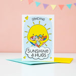 Load image into Gallery viewer, A bright and colourful cute card which features a shining sun hugging a large number of coloured love hearts. The text reads, Sending sunshine and hugs.
