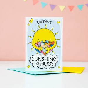 A bright and colourful cute card which features a shining sun hugging a large number of coloured love hearts. The text reads, Sending sunshine and hugs.